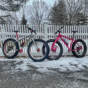 Two fat bikes, one light grey with red components, one hot pink with pink accessories, leaning against a white fence in the snow.