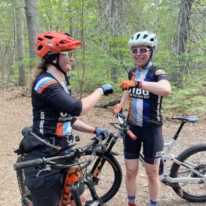 Leslie and Kristin, wearing STBG kits and mountain bike helmets, fist bump in the woods.